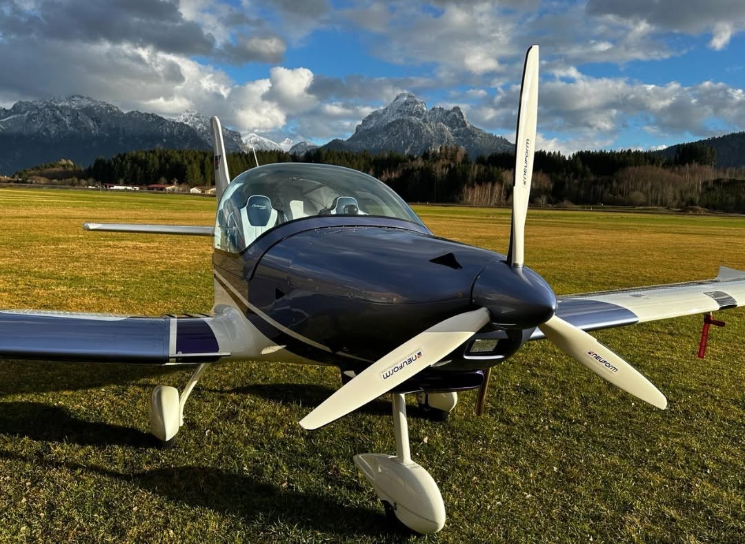 Belmont aircraft parked on an alpine grass strip with snow-capped mountains behind